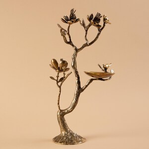May include: A bronze-colored decorative tree sculpture with birds and flowers. The sculpture features a tree trunk base, branches with flowers, and small birds perched on the branches and a birdbath. The overall design is intricate and detailed.