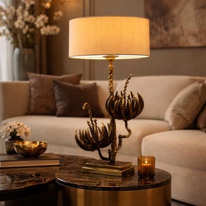 Peut inclure: Lampe de table d&eacute;corative avec abat-jour beige et base florale en bronze. La lampe est pos&eacute;e sur une table ronde &agrave; plateau en marbre fonc&eacute;. Une bougie allum&eacute;e et un bol dor&eacute; sont &eacute;galement sur la table, avec un canap&eacute; beige en arri&egrave;re-plan.