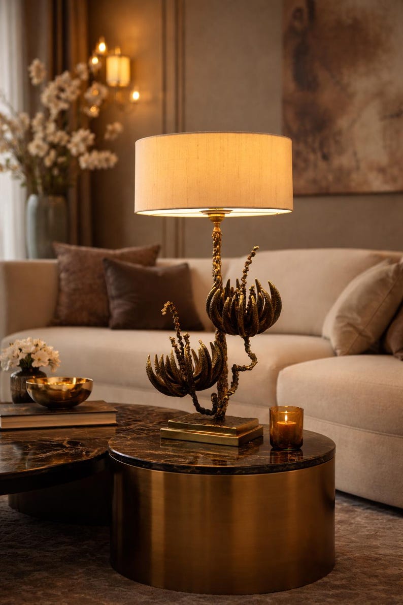 Peut inclure: Lampe de table d&eacute;corative avec abat-jour beige et base florale en bronze. La lampe est pos&eacute;e sur une table ronde &agrave; plateau en marbre fonc&eacute;. Une bougie allum&eacute;e et un bol dor&eacute; sont &eacute;galement sur la table, avec un canap&eacute; beige en arri&egrave;re-plan.