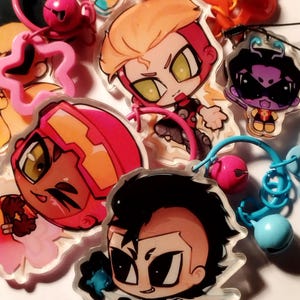 Op de afbeelding: Een verzameling kleurrijke acryl bedels met anime-achtige personages. De bedels hebben verschillende ontwerpen, waaronder een ster en figuren met oranje, rood en zwart haar. Elke bedel heeft een metalen ring voor bevestiging.