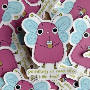 May include: A collection of stickers featuring a cartoonish pink creature with large eyes, blue butterfly wings, and small green boots. The creature holds a coffee cup and the text "perpetually in need of a little treat" is included.
