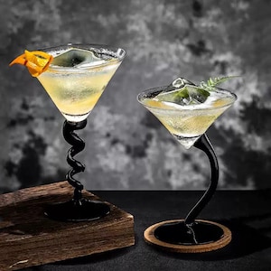 Puede incluir: Dos copas de martini llenas de un cóctel amarillo y hielo. Una copa tiene un tallo en espiral negro, la otra tiene un tallo curvo. Las guarniciones incluyen una cáscara de naranja y una ramita de vegetación. Las copas están sobre un bloque de madera y un posavasos redondo.
