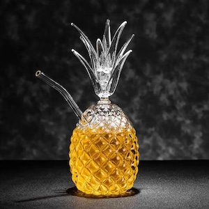 Może przedstawiać: Szklana fajka wodna w kształcie ananasa. Korpus ma wzór w romby i jest wypełniony żółtym płynem. Górna część to korona ananasa z przezroczystego szkła, a zakrzywiona szklana rurka wystaje z boku.