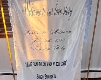 May include: A white fabric wedding banner with the text "Welcome to our love story" and names, date, and location. The banner is held by a gold metal stand and trimmed with blue fabric.