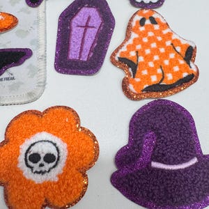 Op de afbeelding: Een verzameling Halloween-thema vilt patches. Ontwerpen omvatten een spook, schedel, kist, heksenhoed en een bloem met een schedel. De patches hebben oranje, paarse, witte en zwarte kleuren met glitteraccenten.