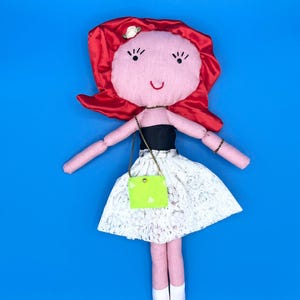 May include: A handmade doll with a pink face, red hair, and a white lace skirt. The doll has a small green purse with a gold chain and is set against a bright blue background. The doll's arms and legs are pink.