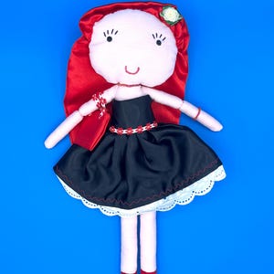 May include: A handmade doll with a light pink face, red hair, and a black dress. The doll has a white flower on its head, a red purse, and red shoes. The dress has a white scalloped hem and a red and white floral belt.