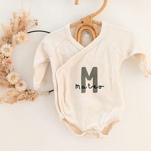Könnte beinhalten: Cremefarbener Baby-Body mit langen Ärmeln, mit dem Namen "Mateo" in einem dunkelgrünen Buchstaben "M" und Schriftzug. Der Body hängt an einem gewebten Holzbügel. Links hängt ein Trockenblumenkranz.