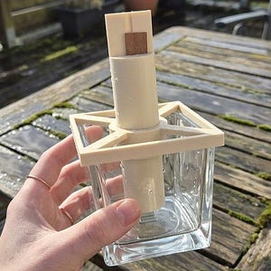 Puede incluir: Un recipiente cuadrado de vidrio transparente con una estructura de plástico beige y un cilindro beige en el interior. Un pequeño trozo de madera es visible en la parte superior del cilindro. El recipiente está sostenido por una mano.