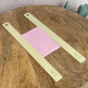 May include: Two brass-colored, rectangular tools with a hole at one end and the number "2 mm" printed on them. They are positioned on a wooden surface, framing a pink rectangular piece. The tools appear to be for measuring or crafting.
