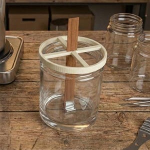Può includere: Un'attrezzatura per la produzione di candele con una pentola in acciaio inossidabile su un fornello, barattoli di vetro e stoppini di legno. Un barattolo ha un supporto per stoppino bianco e beige. Il tavolo è di legno.