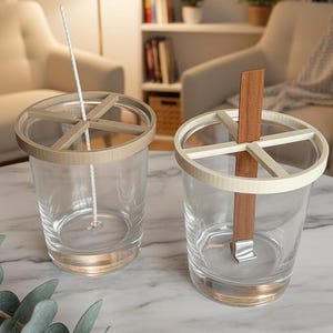 Puede incluir: Dos tarros de velas de vidrio transparente con soportes beige y blancos en forma de cruz. Uno tiene una mecha blanca, el otro una mecha de madera. Los tarros están sobre una superficie de mármol, lo que sugiere un tema de decoración del hogar.