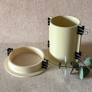 Puede incluir: Dos moldes cilíndricos beige con pinzas negras adjuntas, uno en posición vertical y otro de lado. Los moldes están sobre una superficie texturizada con hojas verdes. Los moldes son probablemente para manualidades o proyectos de bricolaje.