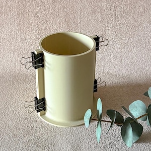 Puede incluir: Un molde cilíndrico verde claro con clips negros que aseguran los lados. El molde se asienta sobre una base circular. Una ramita de hojas verdes está en primer plano, sobre un fondo rosa claro texturizado.