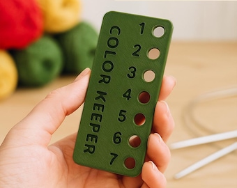 May include: A green Color Keeper tool for knitting, with numbers 1-7 and corresponding holes. The text "COLOR KEEPER" is printed vertically. The background includes yarn balls in red, yellow, and green.