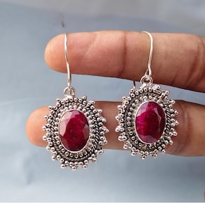 May include: A pair of silver-toned dangle earrings. Each earring features a large, oval-shaped, deep red gemstone, surrounded by a detailed, beaded frame. The earrings are suspended from simple, curved hooks, showcasing a classic design.
