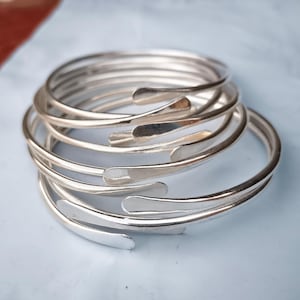Puede incluir: Un conjunto de brazaletes de metal plateado. Cada brazalete presenta un diseño liso y curvado con una pequeña abertura. Los brazaletes están apilados, creando un efecto de capas. El fondo es de color azul claro.