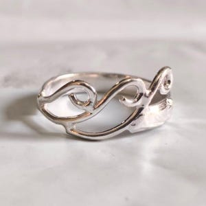 May include: A silver ring with an openwork design featuring swirling, abstract shapes. The ring is set against a light background, highlighting its intricate details and polished finish. This piece of jewelry is suitable for everyday wear.