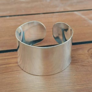 May include: A wide, polished silver-tone cuff bracelet with a smooth, reflective surface and a prominent open gap at the back. The metal band features a clean, curved silhouette with rounded edges, showcasing a bright, metallic finish throughout.