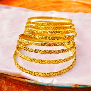 Op de afbeelding: Een stapel gouden armbanden met ingewikkelde geometrische en bloemmotieven. De armbanden zijn gerangschikt op een wit oppervlak, waarbij hun gedetailleerde ontwerpen en stralende glans worden benadrukt. De sieraden zijn een klassiek accessoire.