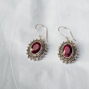 Peut inclure: Une paire de boucles d'oreilles pendantes en argent. Chaque boucle d'oreille présente une grosse pierre précieuse ovale rouge foncé, sertie dans un cadre argenté détaillé en forme d'éclat de soleil. Les boucles ont une fermeture à crochet simple.