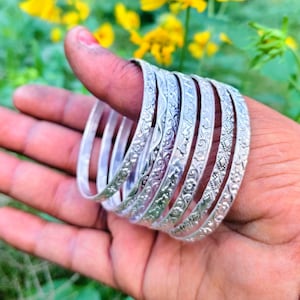 Puede incluir: Una mano muestra una colección de pulseras de plata. Cada pulsera presenta intrincados diseños florales y geométricos. Las pulseras están apiladas en el pulgar y sostenidas en la palma, mostrando su diseño contra un fondo borroso de follaje verde y flores amarillas.
