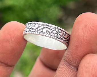 May include: A silver ring with a detailed floral and scrollwork design. The ring's surface is adorned with repeating stylized flowers and curved lines, creating a textured appearance. The ring is held between two fingers, highlighting its intricate craftsmanship.