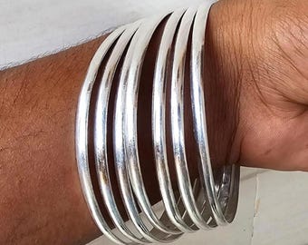 Peut inclure: Un ensemble de sept bracelets de couleur argentée est empilé sur un poignet. Les bracelets sont lisses et brillants, reflétant la lumière. Le bijou est un accessoire simple et élégant.