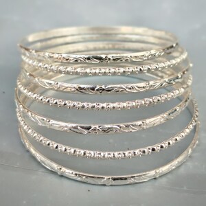 May include: A stack of seven silver-toned bangles. The bracelets feature alternating designs, with some bands displaying a smooth, engraved floral pattern and others showcasing a textured, beaded surface with small, raised metallic dots.