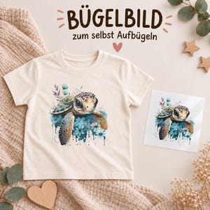 Könnte beinhalten: Ein cremefarbenes Kurzarm-T-Shirt mit einer Aquarell-Grafik einer Meeresschildkröte, umgeben von blauen Blumen und Blättern. Neben dem T-Shirt liegt ein quadratischer Transferbogen, der dieselbe Illustration der Meeresschildkröte zeigt.