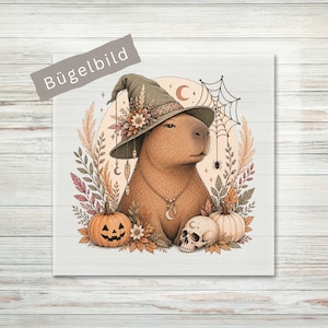 Könnte beinhalten: Ein quadratisches Bügelbild mit einem braunen Capybara, das einen mit Blumen verzierten grünen Hexenhut trägt. Die Illustration zeigt einen Kürbis, einen Totenkopf, Herbstblätter, ein Spinnennetz und eine Mondsichel. Der Text Bügelbild ist zu sehen.
