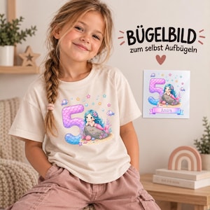 Könnte beinhalten: Ein cremefarbenes T-Shirt mit einer Grafik einer großen lila Zahl fünf, einer Meerjungfrau, Fischen und Sternen. Ein quadratischer Bügeltransfer zeigt dasselbe Motiv mit dem Namen Amira und dem Text BÜGELBILD zum selbst Aufbügeln.