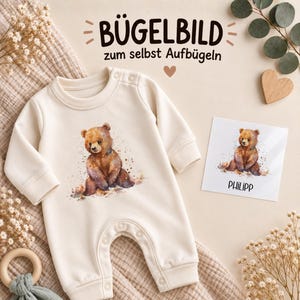 Könnte beinhalten: Ein cremefarbener langärmeliger Baby-Strampler mit einer aquarellartigen Illustration eines braunen Bären auf der Brust. Daneben liegt eine quadratische Karte, die dasselbe Bärenmotiv über dem Namen PHILIPP in schwarzen Großbuchstaben zeigt.