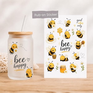 Könnte beinhalten: Ein mattiertes Glas mit Bambusdeckel und transparentem Strohhalm, verziert mit einer Biene und dem Schriftzug "bee happy". Daneben liegt ein Bogen mit Rub-on-Stickern, die Bienen, Honigtöpfe sowie die Texte "good vibes only" und "bee happy" zeigen.