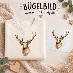 Könnte beinhalten: Ein cremefarbenes Sweatshirt mit einer aquarellartigen Illustration eines Hirschkopfes mit großem Geweih auf der Vorderseite, begleitet von einem separaten quadratischen Transferblatt, das dasselbe Hirschmotiv und den Text BÜGELBILD zum selbst Aufbügeln zeigt.