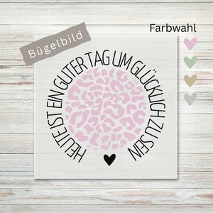 Könnte beinhalten: Ein weißes Bügelbild mit einem zentralen hellrosa Leopardenmuster-Kreis, umgeben von dem schwarzen Text "HEUTE IST EIN GUTER TAG UM GLÜCKLICH ZU SEIN" und einem kleinen schwarzen Herz unten, mit Farbauswahl in Form von vier kleinen Herzen.