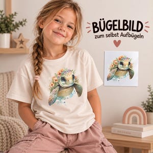 Könnte beinhalten: Ein cremefarbenes T-Shirt mit einer Grafik einer Meeresschildkröte, die mit bunten Blumen auf dem Kopf geschmückt ist. Daneben liegt ein quadratischer Bügeltransfer mit demselben Schildkrötenmotiv und dem Text BÜGELBILD zum selbst Aufbügeln.