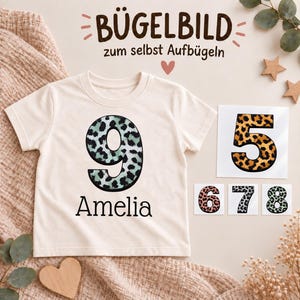 Könnte beinhalten: Ein cremefarbenes Kurzarm-T-Shirt mit einer großen Neun im blaugrün-schwarzen Leopardenmuster und dem Namen Amelia darunter. Enthalten sind vier separate Bügelbilder mit den Zahlen fünf, sechs, sieben und acht für den eigenen Gebrauch.