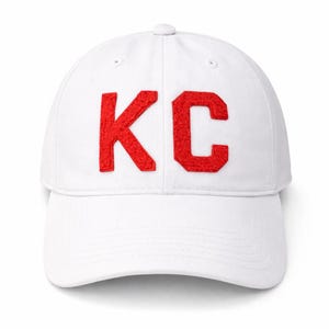 May include: White baseball cap with the red letters "KC" embroidered on the front. The cap has a curved brim and an adjustable strap at the back. A classic, casual accessory.