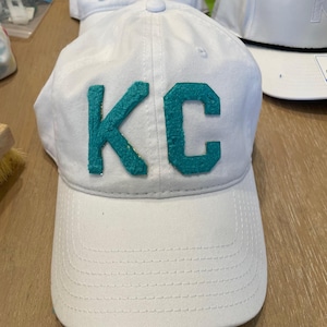May include: White baseball cap with teal "KC" letters. The cap has a curved brim and a button on top. Other white caps with different colored letters are visible in the background.
