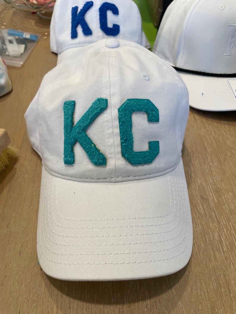 May include: White baseball cap with teal "KC" letters. The cap has a curved brim and a button on top. Other white caps with different colored letters are visible in the background.