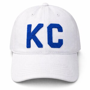 May include: White baseball cap with the blue letters "KC" on the front. The cap has a curved brim and a button on top. The letters are made of a textured material.