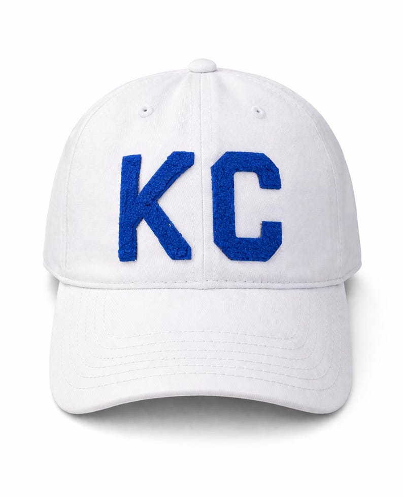 May include: White baseball cap with the blue letters "KC" on the front. The cap has a curved brim and a button on top. The letters are made of a textured material.