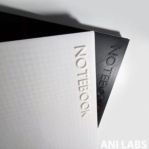 May include: Two notebooks, one white and one black, are stacked at an angle. The word "NOTEBOOK" is embossed on the cover of each notebook. The white notebook has a grid pattern. The bottom right corner of the image has the text "ANI LABS".