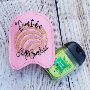 May include: Pink fabric hand sanitizer holder with embroidered text "Don't be Self Conchas" and a pink and gold concha design. The holder is holding a clear plastic bottle of hand sanitizer with a green label that says "Island Margarita".