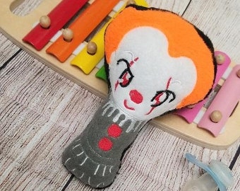 May include: A plush toy shaped like a clown's head with a white face, orange hair, and black eyes. The toy is attached to a gray fabric body with red buttons. The toy is sitting on a wooden xylophone with colorful keys.