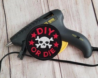 Può includere: Una pistola per colla a caldo nera e gialla con un cavo nero e una toppa ricamata rossa, bianca e nera che dice "DIY or DIE" con un teschio e ossa incrociate al centro.