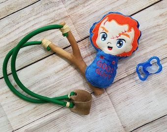 May include: A handmade slingshot with green rubber tubing and a brown leather pouch. Beside it is a plush doll of a character with orange hair and a blue outfit that says "GOOD GUYS". A blue pacifier is also present.