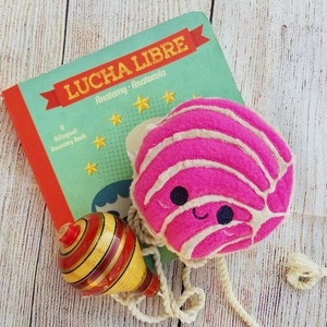 Pu&ograve; includere: Un giocattolo di peluche rosa e bianco a forma di concha, un pane dolce messicano tradizionale. Il giocattolo ha un viso sorridente ed &egrave; attaccato a una corda e una trottola di legno con strisce rosse, gialle e dorate.