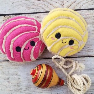 Pu&ograve; includere: Due giocattoli di peluche a forma di pane dolce messicano, uno rosa e uno giallo, con facce sorridenti. Una trottola di legno con strisce rosse e gialle &egrave; in primo piano.
